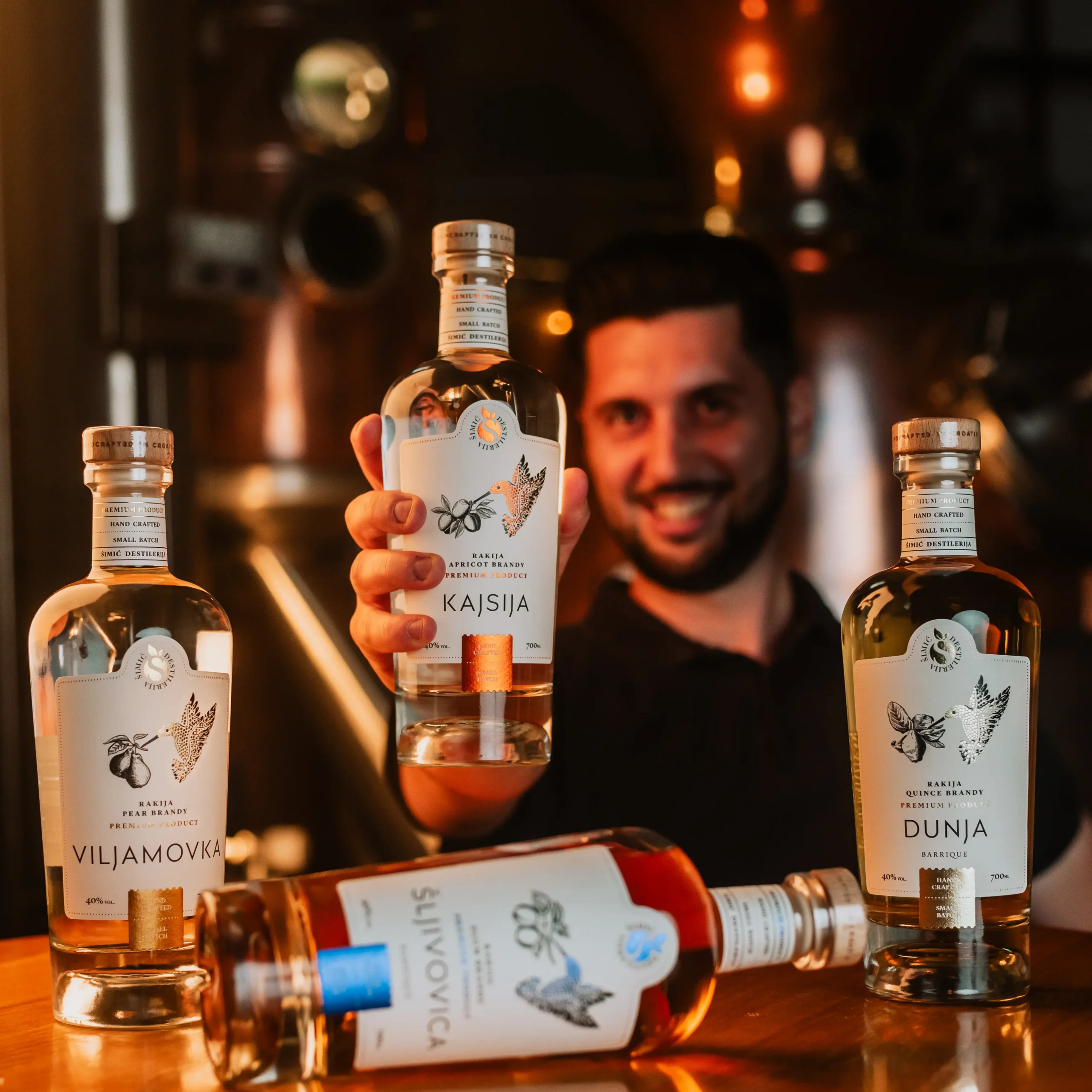 Smiling man holding Kajsija apricot brandy bottle with Viljamovka, Dunja, and Šljivovica rakija bottles